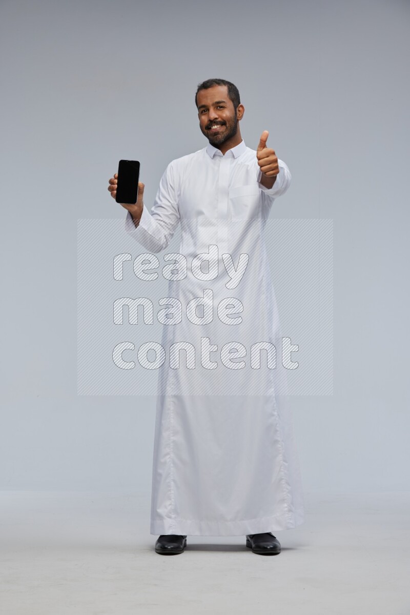 Saudi man wearing Thob standing showing phone to camera on Gray background