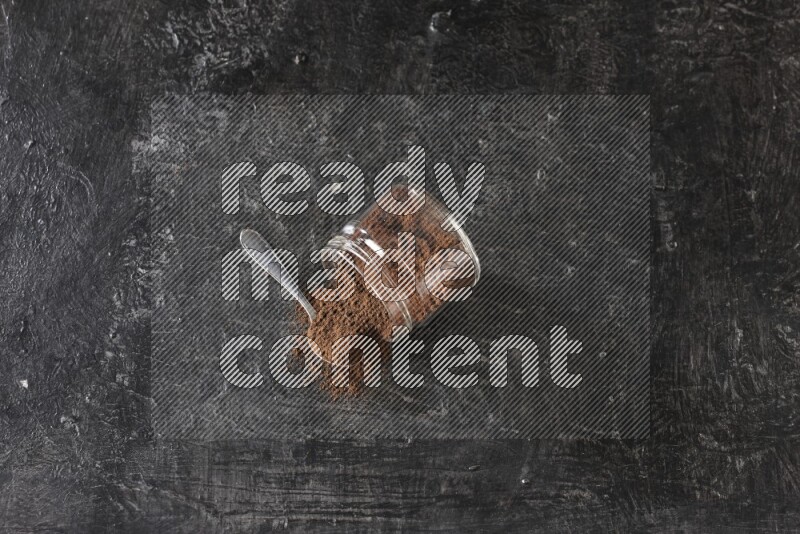A flipped glass jar and metal spoon full of cloves powder with cloves spread on a textured black flooring