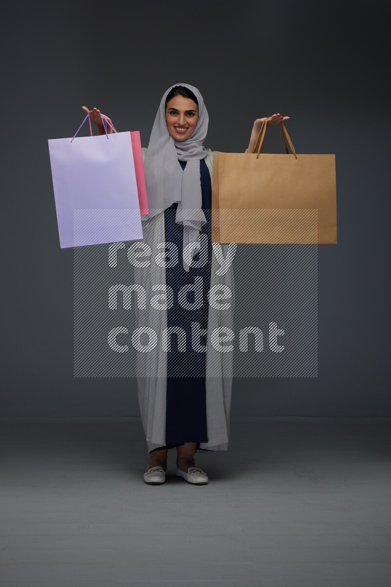 A Saudi woman wearing a light gray Abaya and head scarf standing and holding shopping bags on a grey background