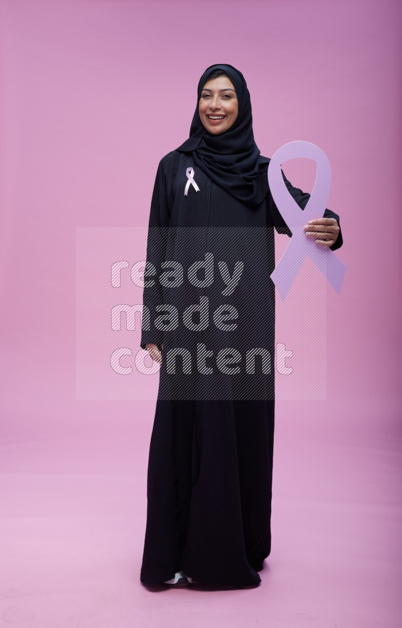 Saudi woman wearing pink ribbon on abaya standing holding awareness ribbon on plain pink background