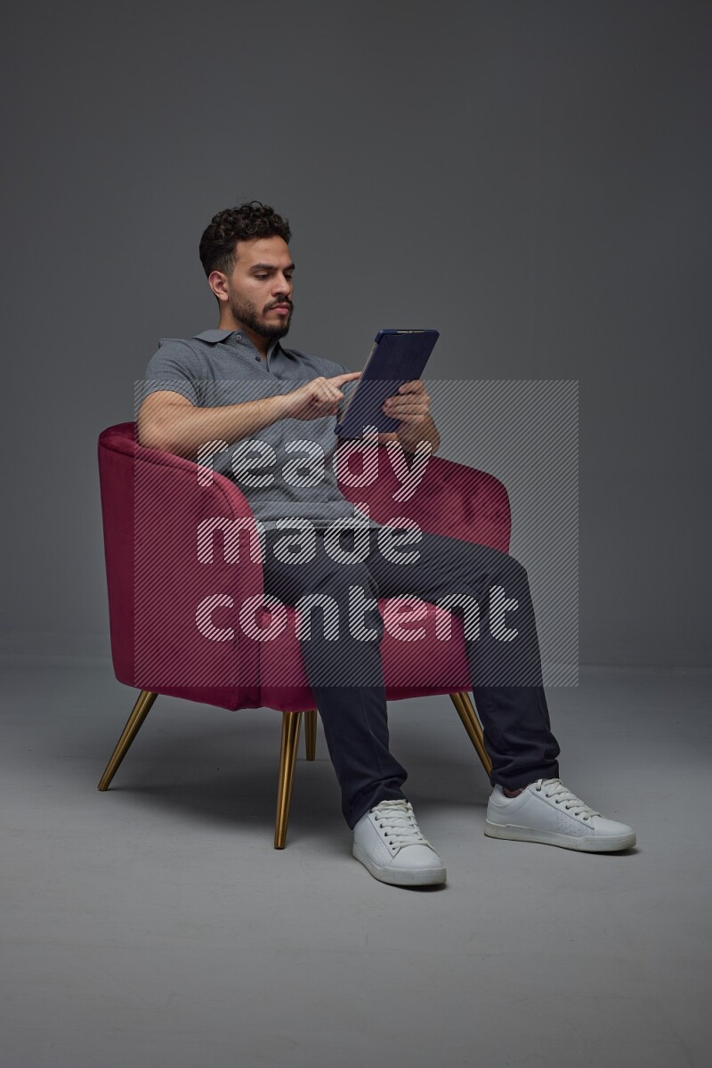 A man wearing casual and using his tablet while sitting on a burgundy chair eye level on a gray background