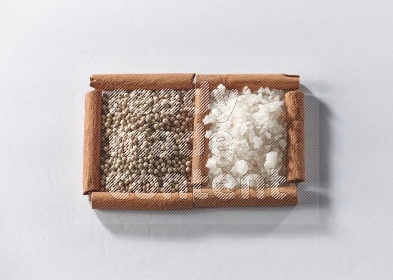 مربعان من أعواد القرفة ممتلئان بحبوب الفلفل الأبيض والملح الأبيض علي خلفية بيضاء