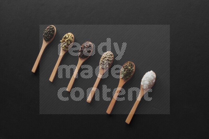 wooden spoons filled with white peppers, cloves, cardamom, salt, black peppers and basil on black flooring