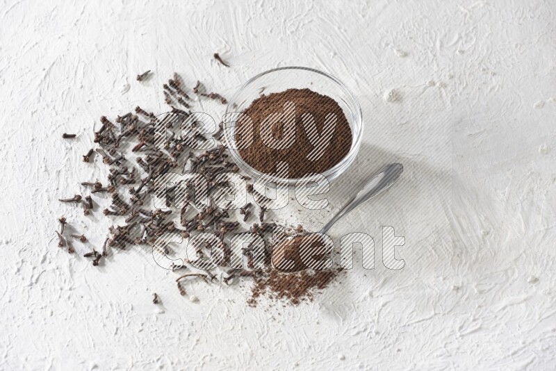 A glass bowl and a metal spoon full of cloves powder with cloves grains spread on a textured white flooring