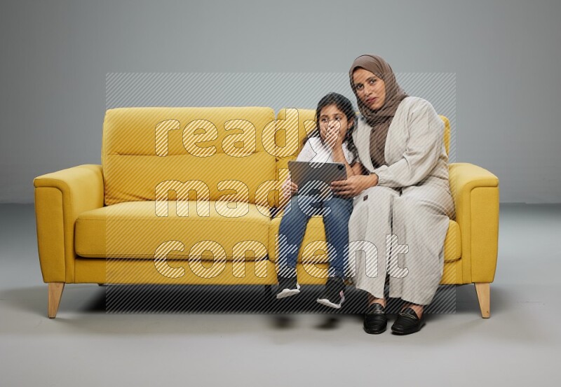 A girl with her mother on yellow sofa and watching on iPad on gray background