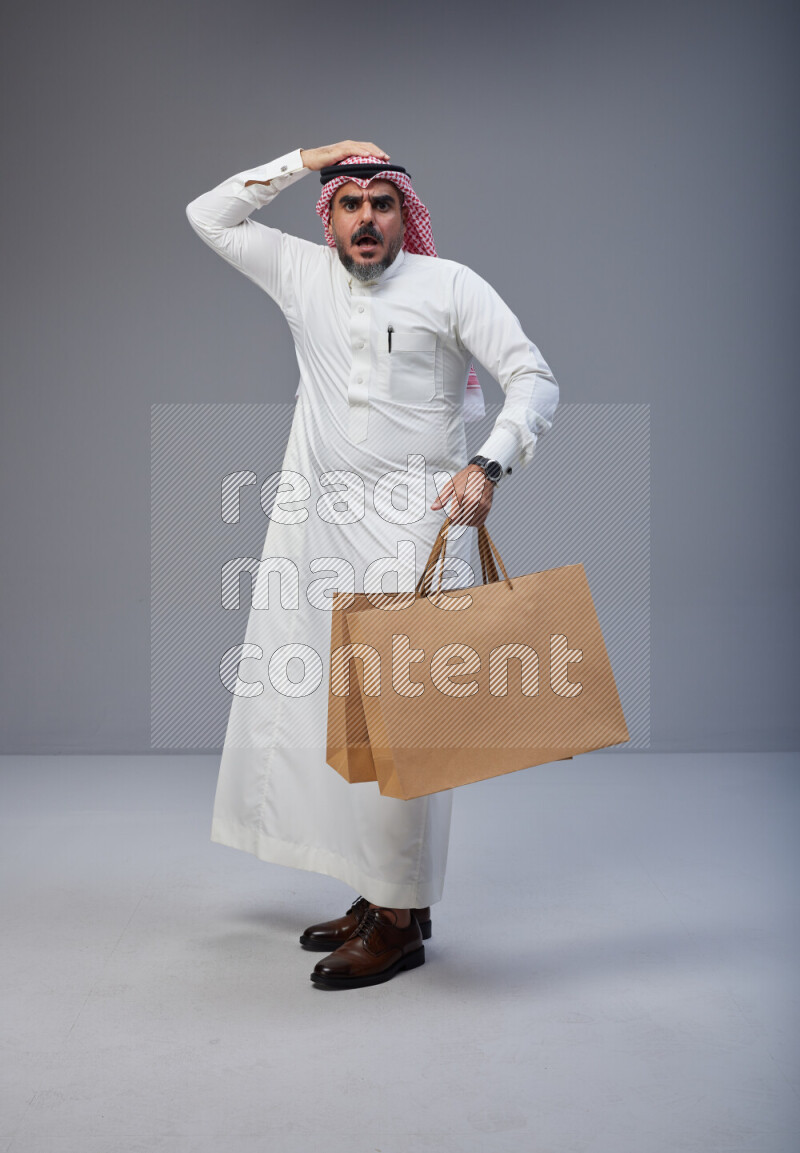 Saudi man Wearing Thob and red Shomag standing holding shopping bag on Gray background