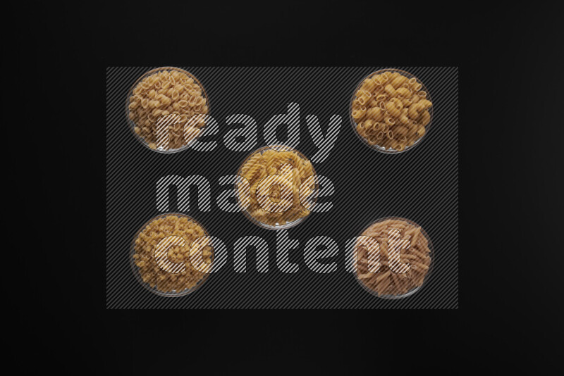 Different pasta types in 5 glass bowls on black background