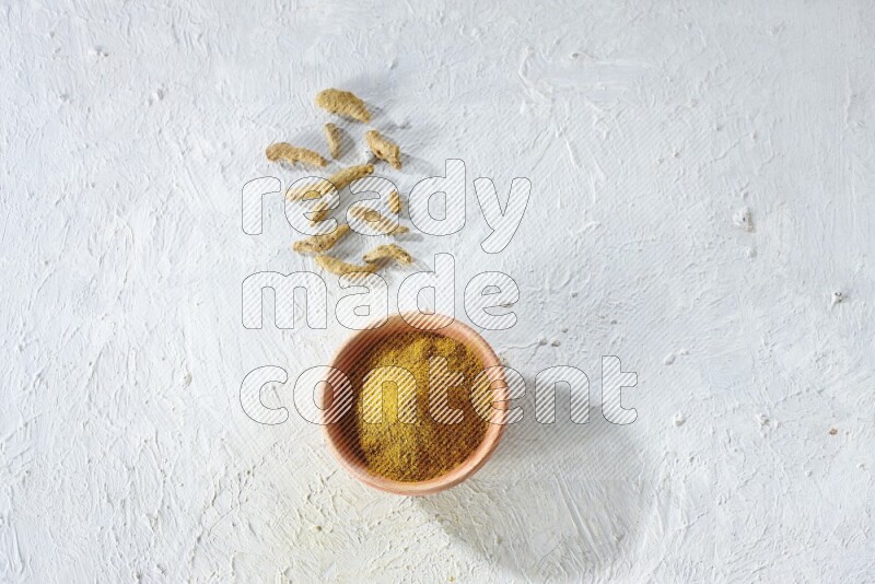 A wooden bowl full of turmeric powder with dried whole fingers on textured white flooring