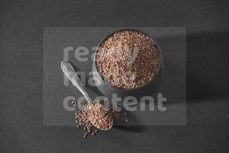 A black pottery bowl full of flaxseeds and a metal spoon full of the seeds on a black flooring