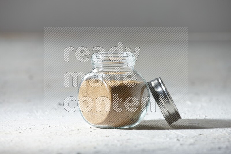 A glass spice jar full of cumin powder on textured white flooring