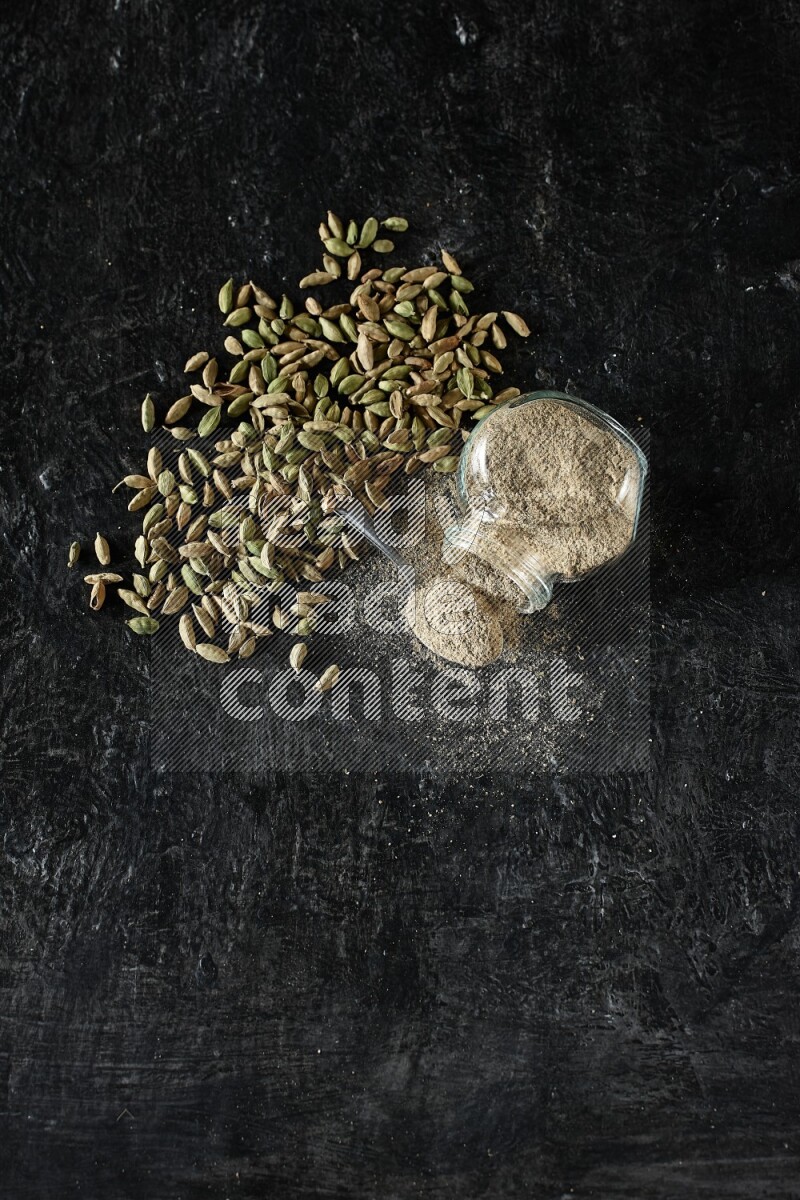 A flipped glass spice jar and a metal spoon full of cardamom powder and cardamom seeds spreaded on textured black flooring
