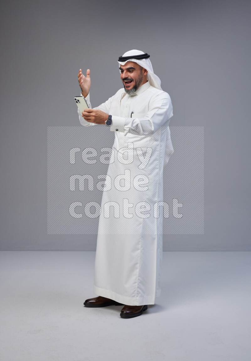 Saudi man Wearing Thob and white Shomag standing texting on phone on Gray background