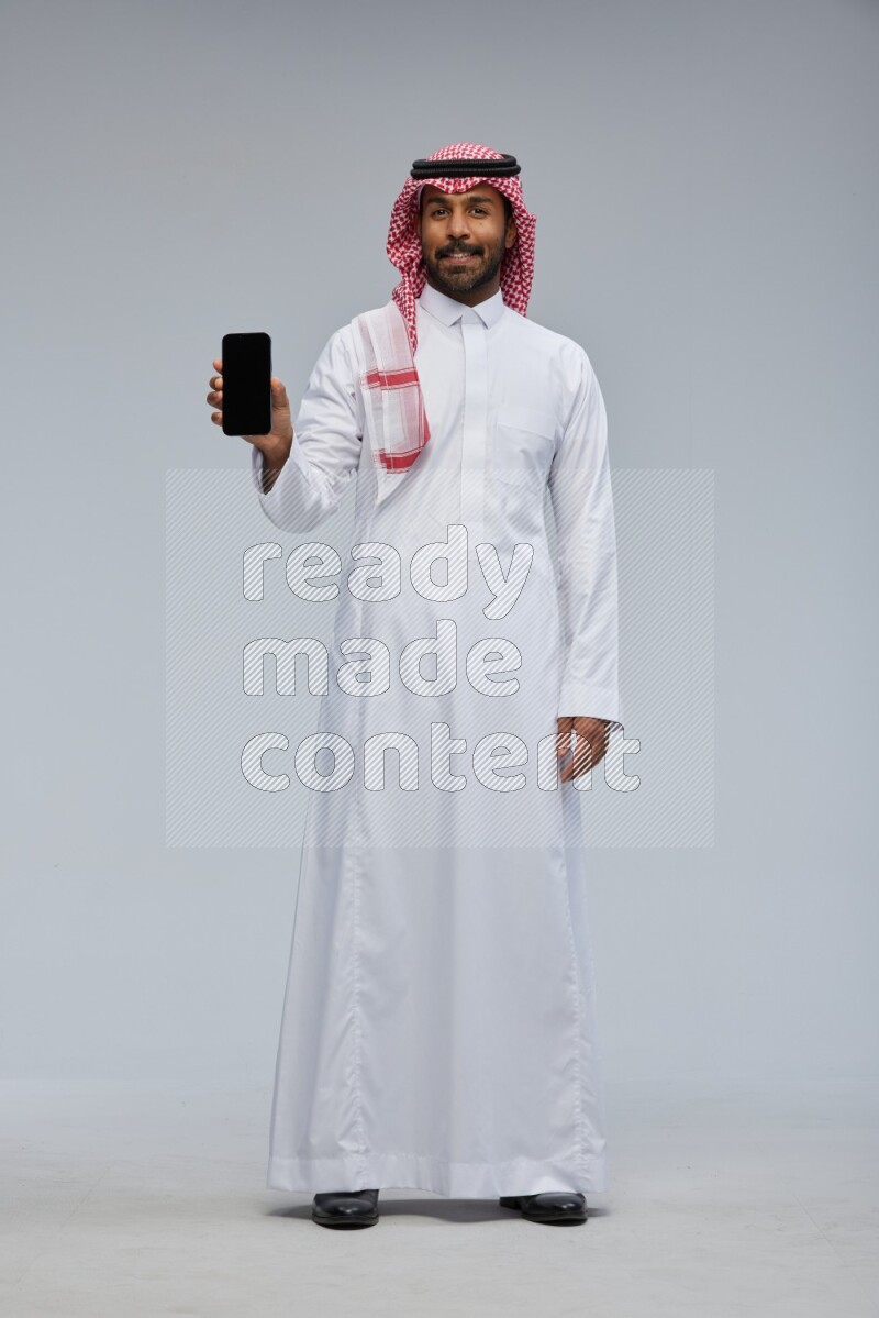 Saudi man Wearing Thob and shomag standing showing phone to camera on Gray background