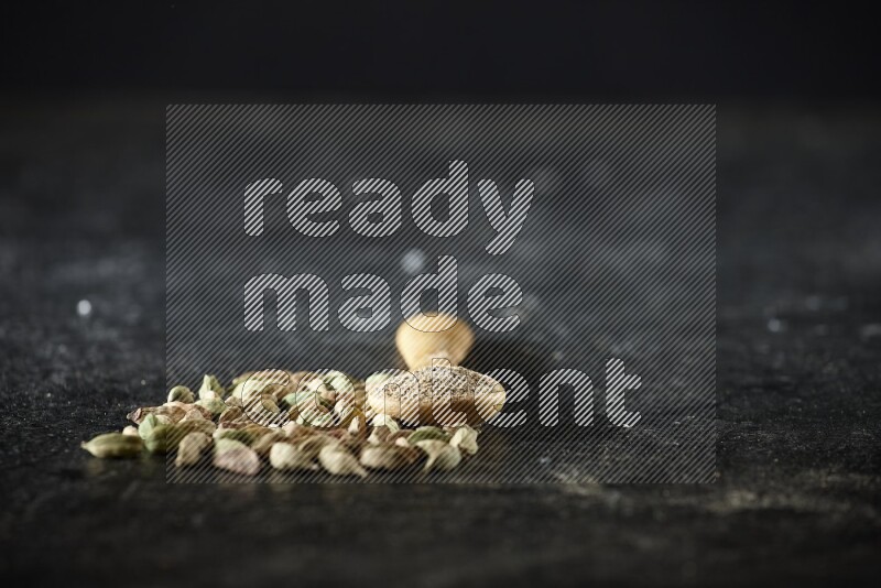 A wooden spoon full of cardamom powder and cardamom seeds next of it on textured black flooring