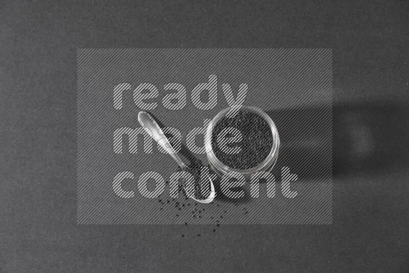 A glass jar full of black seeds with a metal spoon full of the seeds on a black flooring