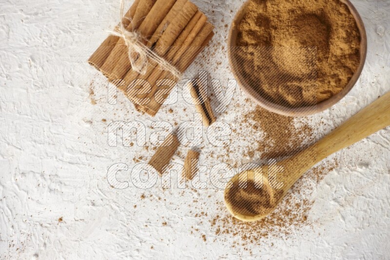 Cinnamon sticks stacked and bounded beside a wooden bowl full of cinnamon powder and a wooden spoon full of powder on white background