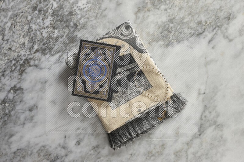 A folded prayer mat with different elements such as quran and prayer beads on grey marble background
