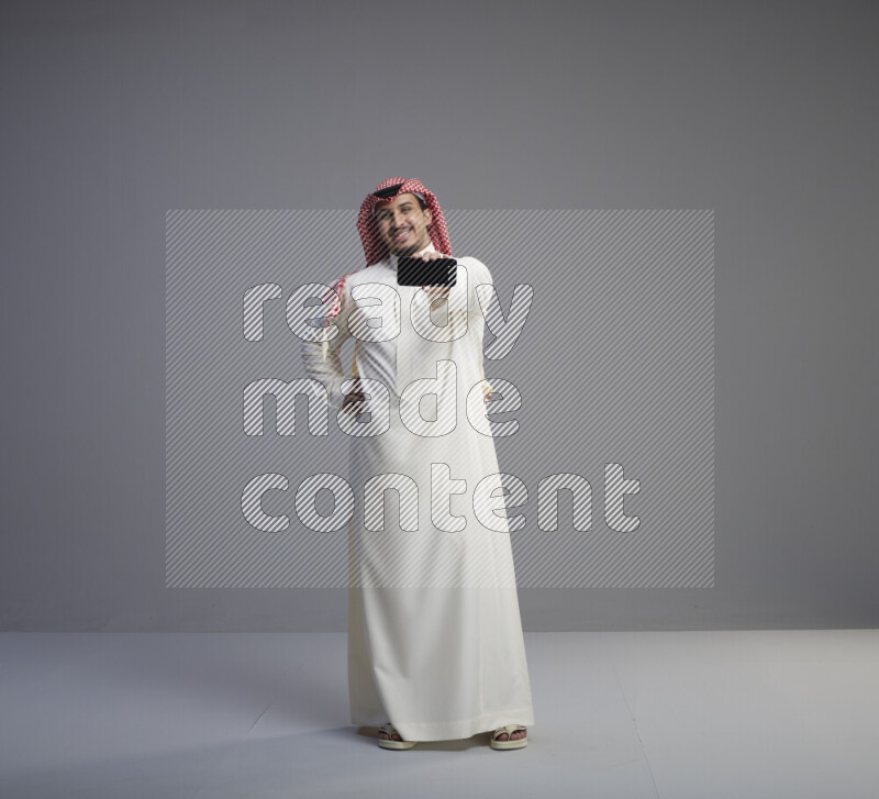 A Saudi man standing wearing thob and red shomag showing phone to camera on gray background