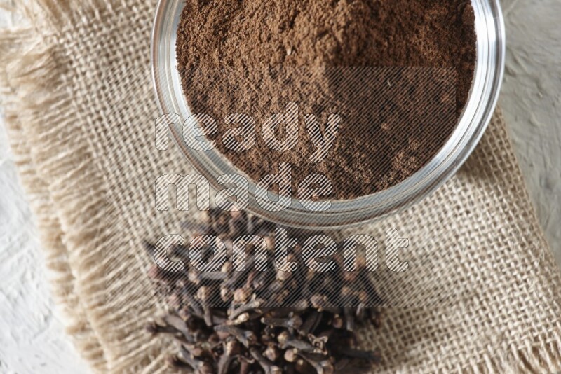 وعاء زجاجي ممتلئ ببودرة القرنفل مع حبوب القرنفل علي قطعة قماش على أرضية بيضاء