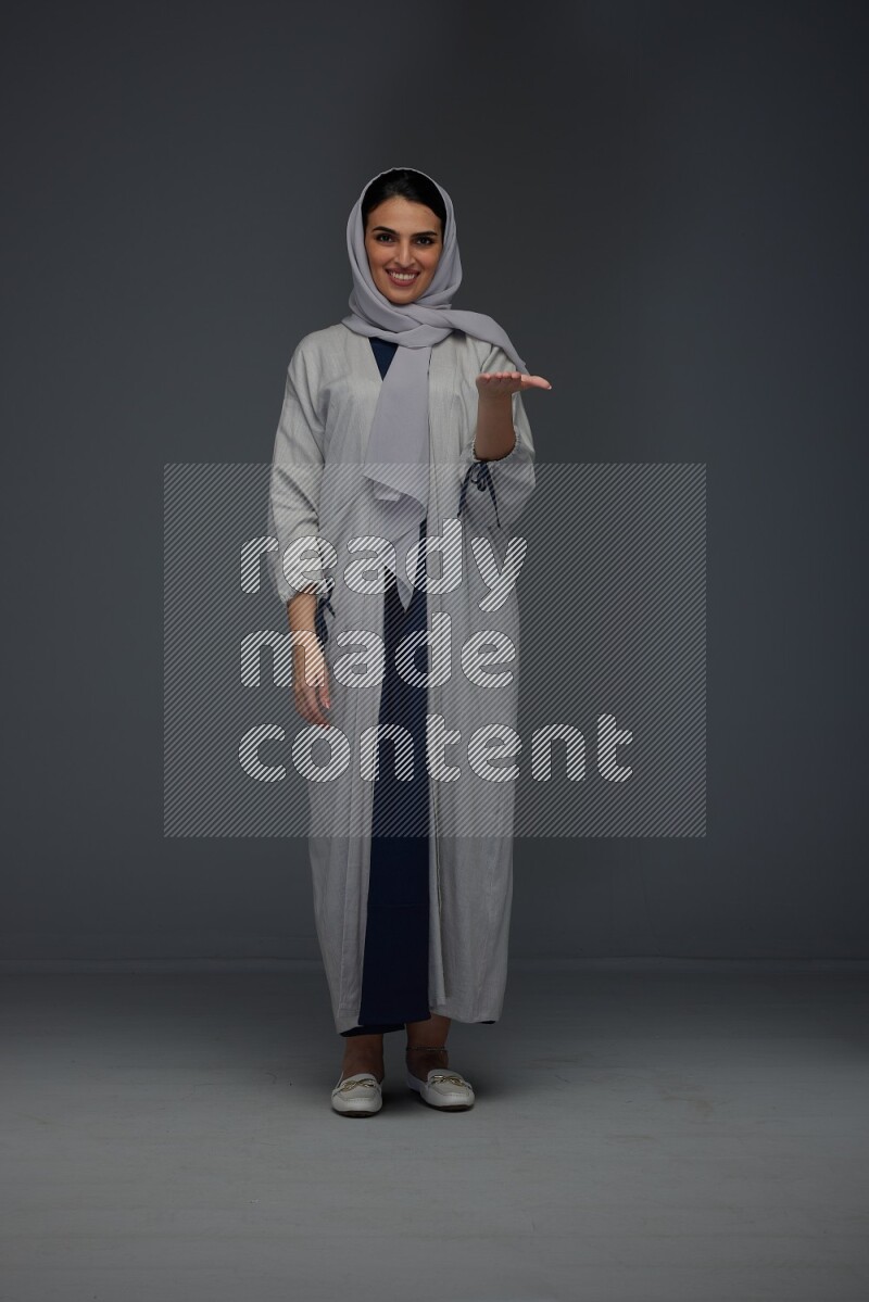 A Saudi woman wearing a light gray Abaya and head scarf standing and pointing in different directions eye level on a grey background