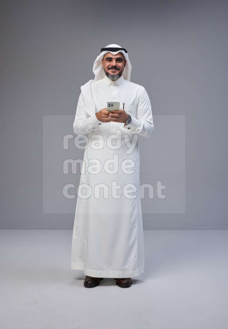 Saudi man Wearing Thob and white Shomag standing texting on phone on Gray background