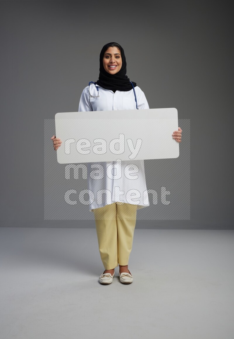 Saudi woman wearing lab coat with stethoscope standing holding board on Gray background