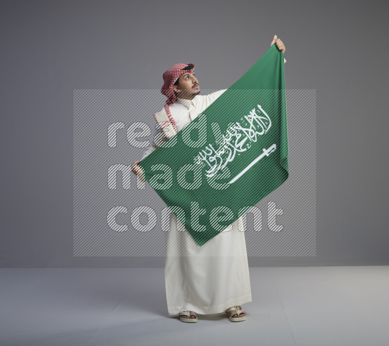 A saudi man standing wearing thob and red shomag holding big saudi flag on gray background