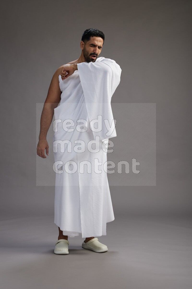 A man wearing Ehram standing showing how to sneeze on gray background