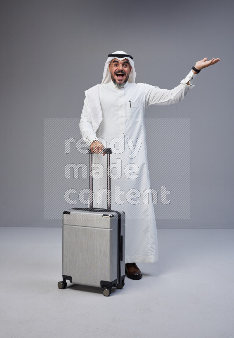 Saudi man wearing Thob and white Shomag standing holding Travel bag on Gray background