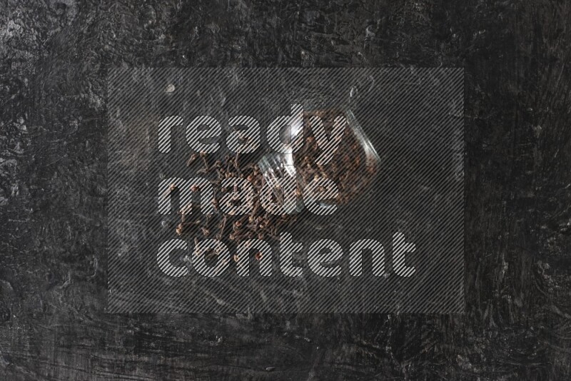 A flipped glass spice jar full of cloves and cloves came out of it on textured black flooring
