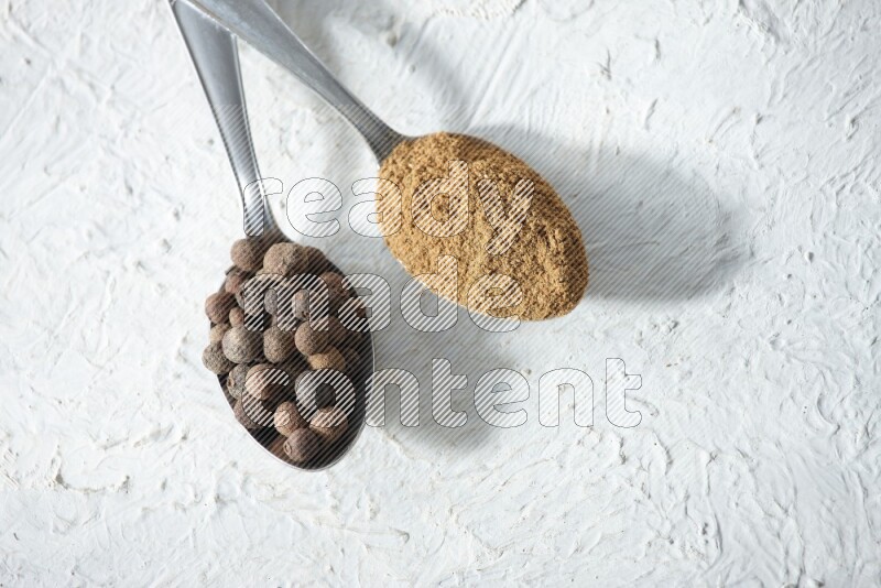 2 metal spoons full of allspice powder and whole balls on a textured white flooring