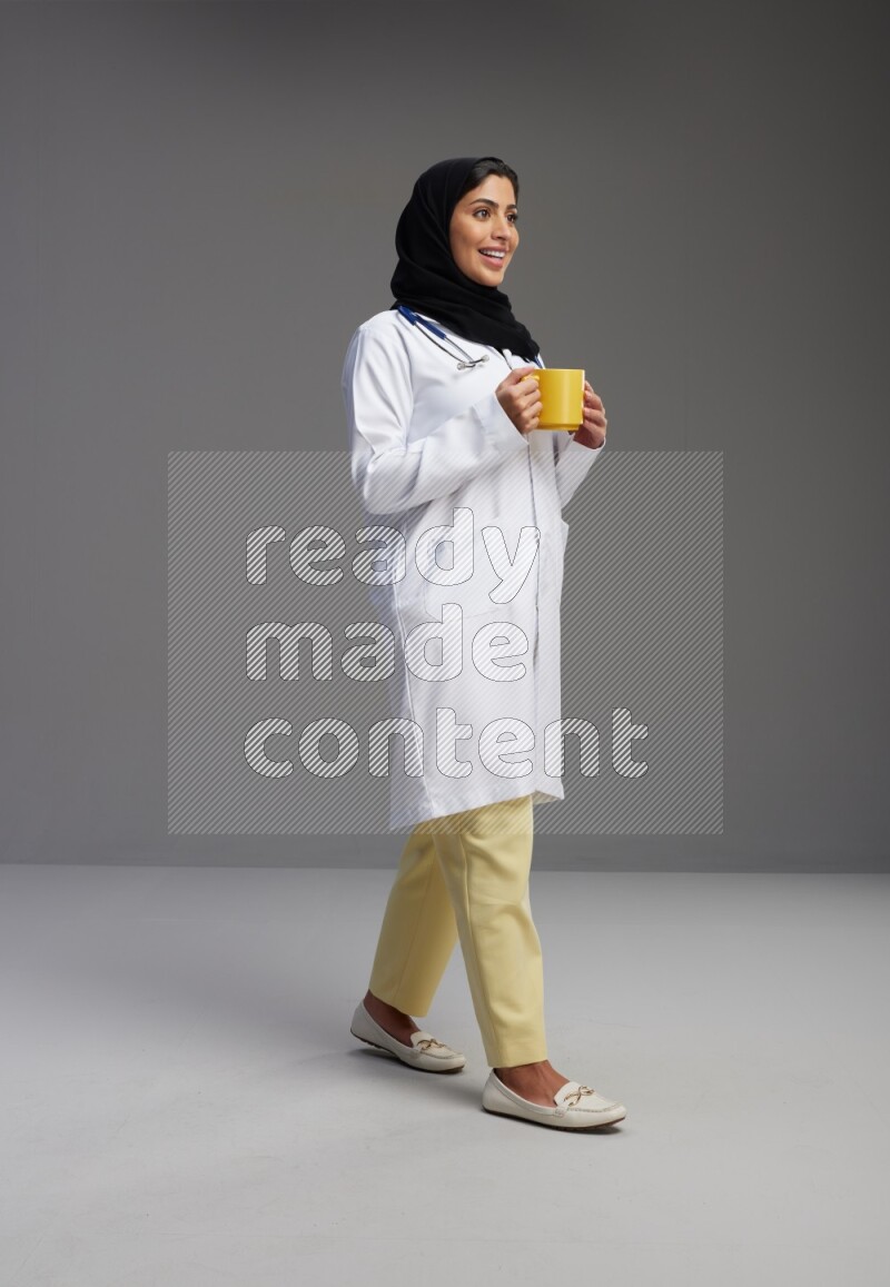 Saudi woman wearing lab coat with stethoscope standing holding mug on Gray background