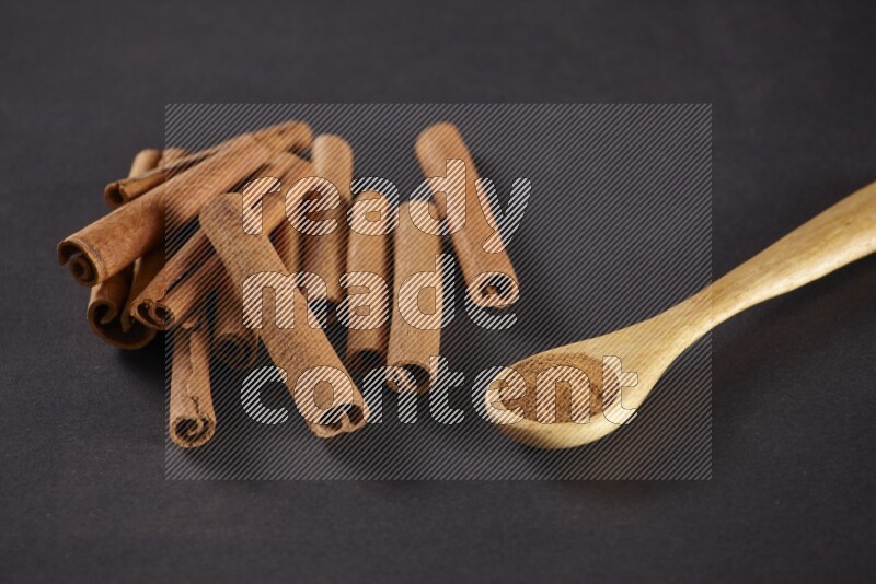 Cinnamon sticks stacked beside a wooden spoon full of cinnamon powder on black background