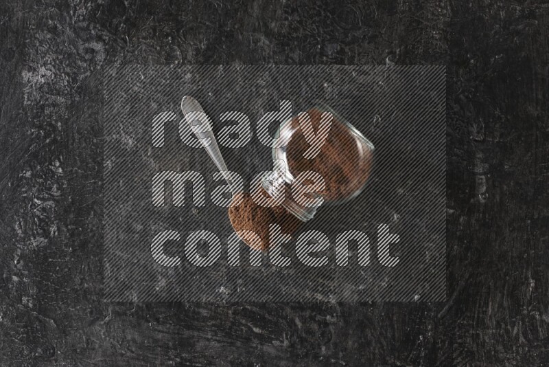 A flipped glass spice jar and a metal spoon full of cloves powder on textured black flooring