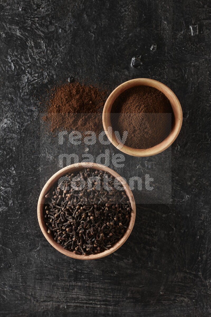 2 wooden bowls full of cloves powder and whole cloves on a textured black flooring