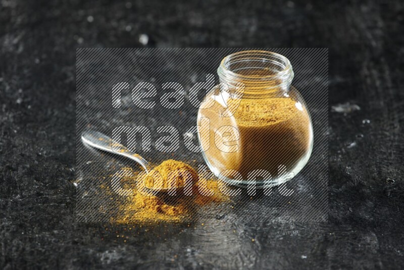 A glass spice jar and metal spoon full of turmeric powder on textured black flooring