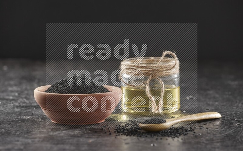 A wooden bowl and spoon full of black seeds and a glass jar of black seeds oil on a textured black flooring