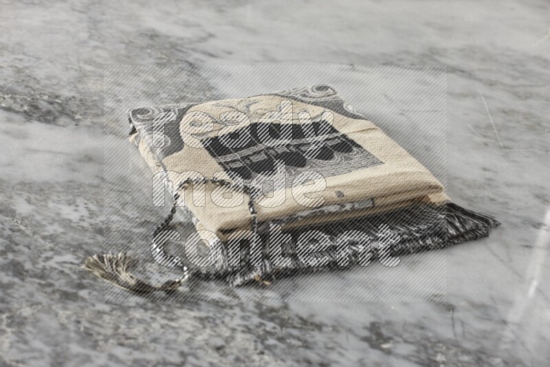 A folded prayer mat with different elements such as quran and prayer beads on grey marble background
