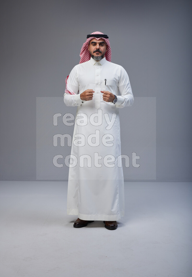 Saudi man Wearing Thob and red white Shomag standing interacting with the camera on Gray background