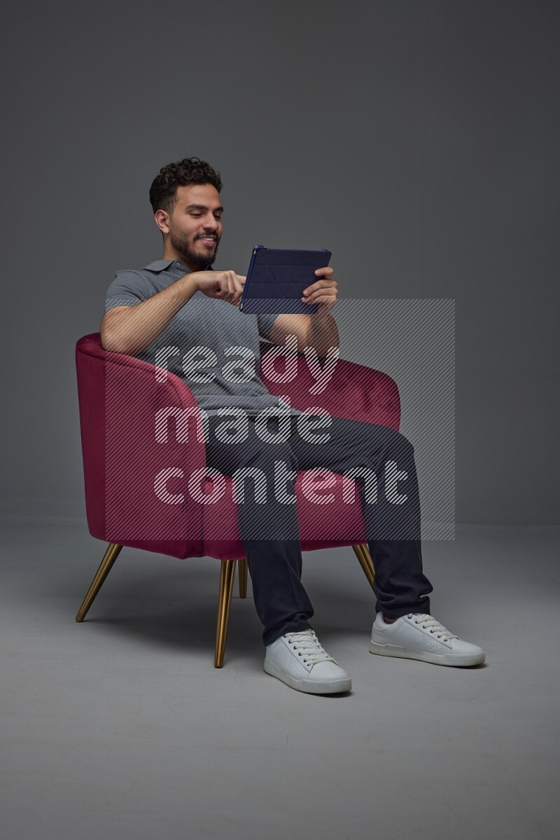 A man wearing casual and using his tablet while sitting on a burgundy chair eye level on a gray background