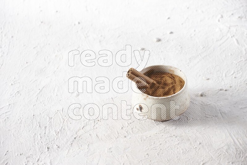 Ceramic beige bowl full of cinnamon powder with a cinnamon stick on a textured white background