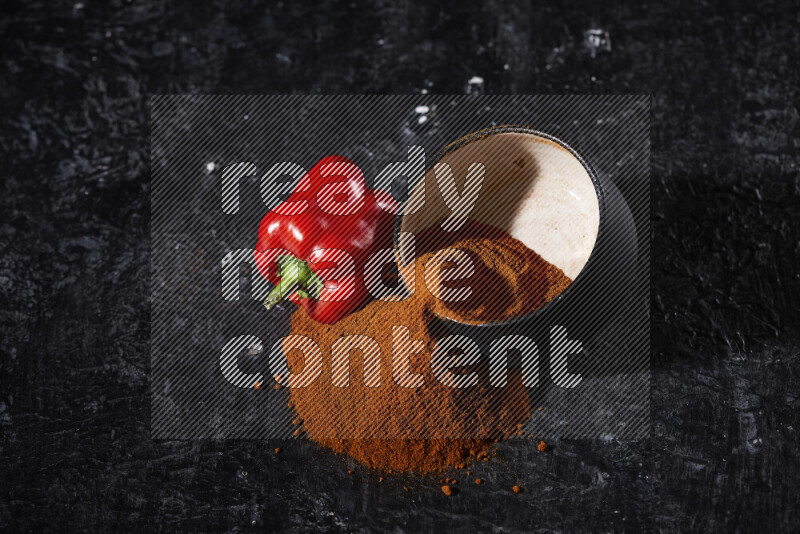 A black pottery bowl full of ground paprika powder with fallen powder from it on black background