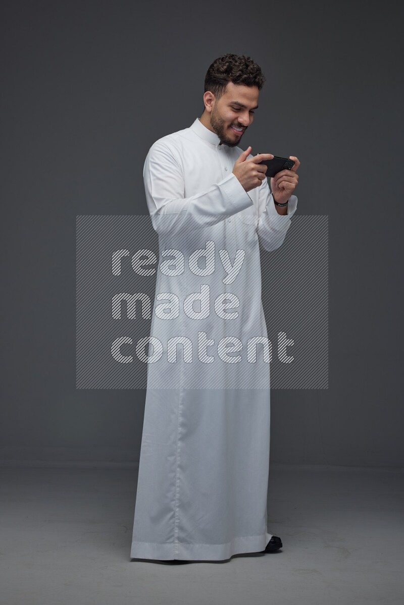A Saudi man wearing Thobe and playing using his phone horizontally playing video games while standing and making different poses eye level on a gray background
