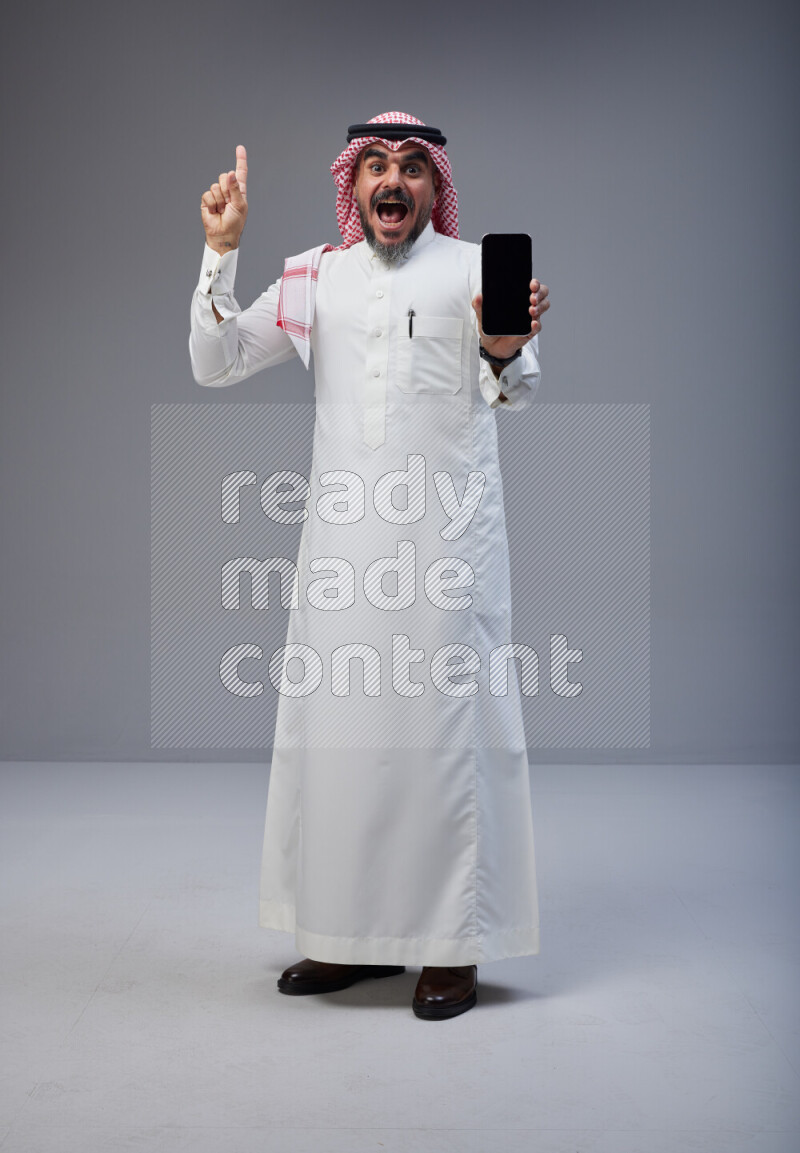 Saudi man Wearing Thob and red Shomag standing showing phone to camera on Gray background