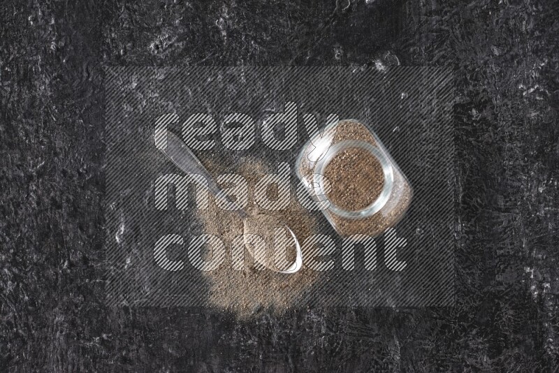 A glass spice jar full of black pepper powder and a metal spoon full of powder on textured black flooring