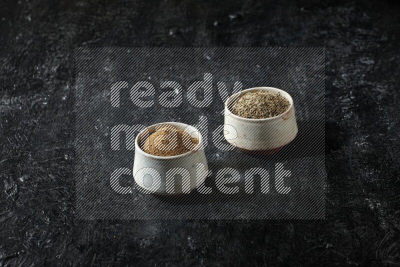 2 beige bowls, one full of cumin powder and the other full of seeds on a textured black flooring
