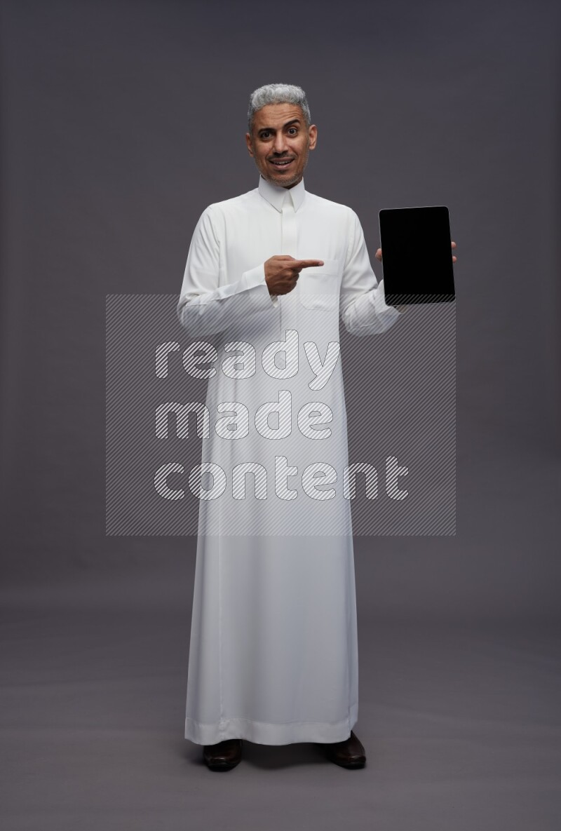 Saudi man wearing thob standing showing tablet to camera on gray background