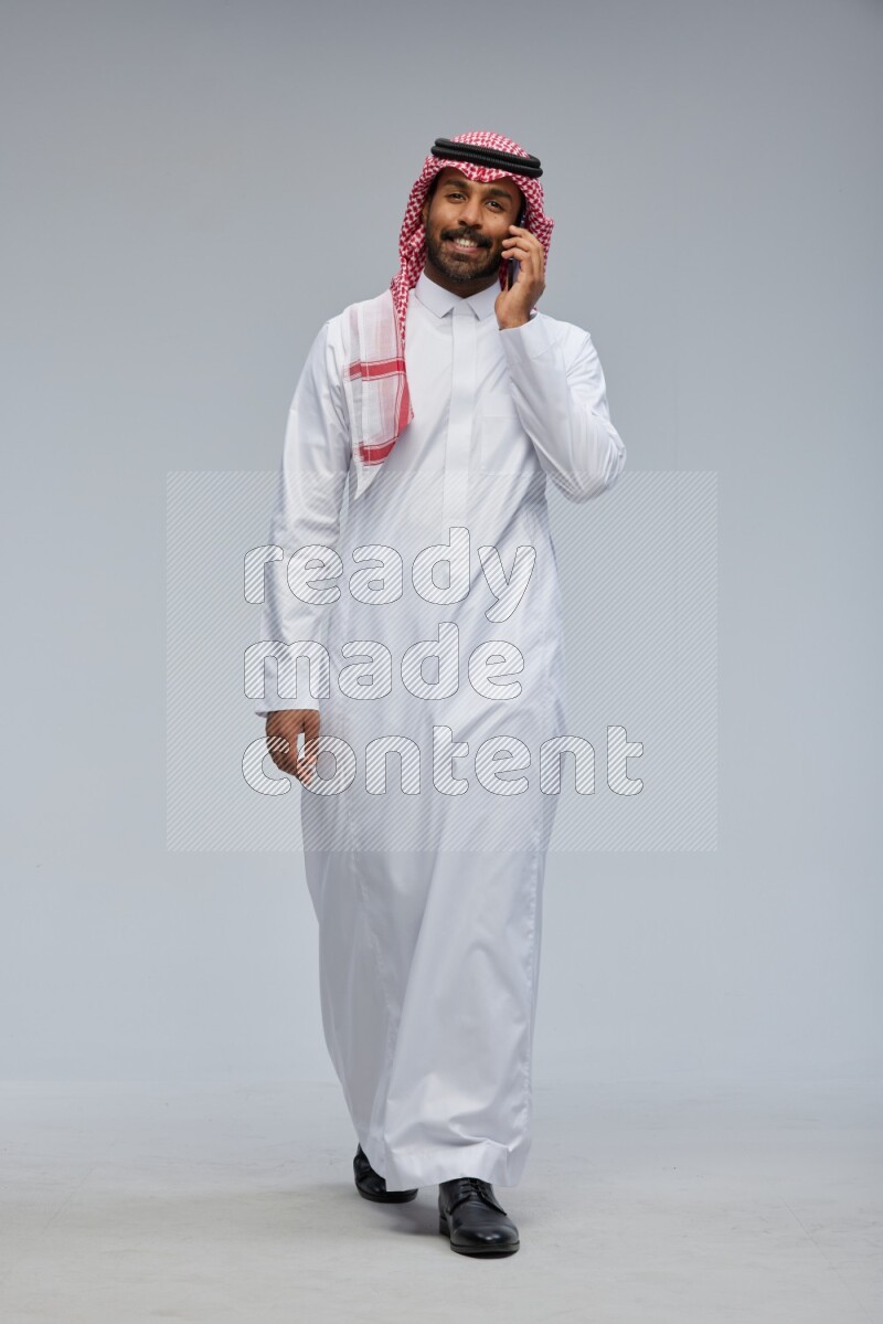Saudi man Wearing Thob and shomag standing talking on phone on Gray background