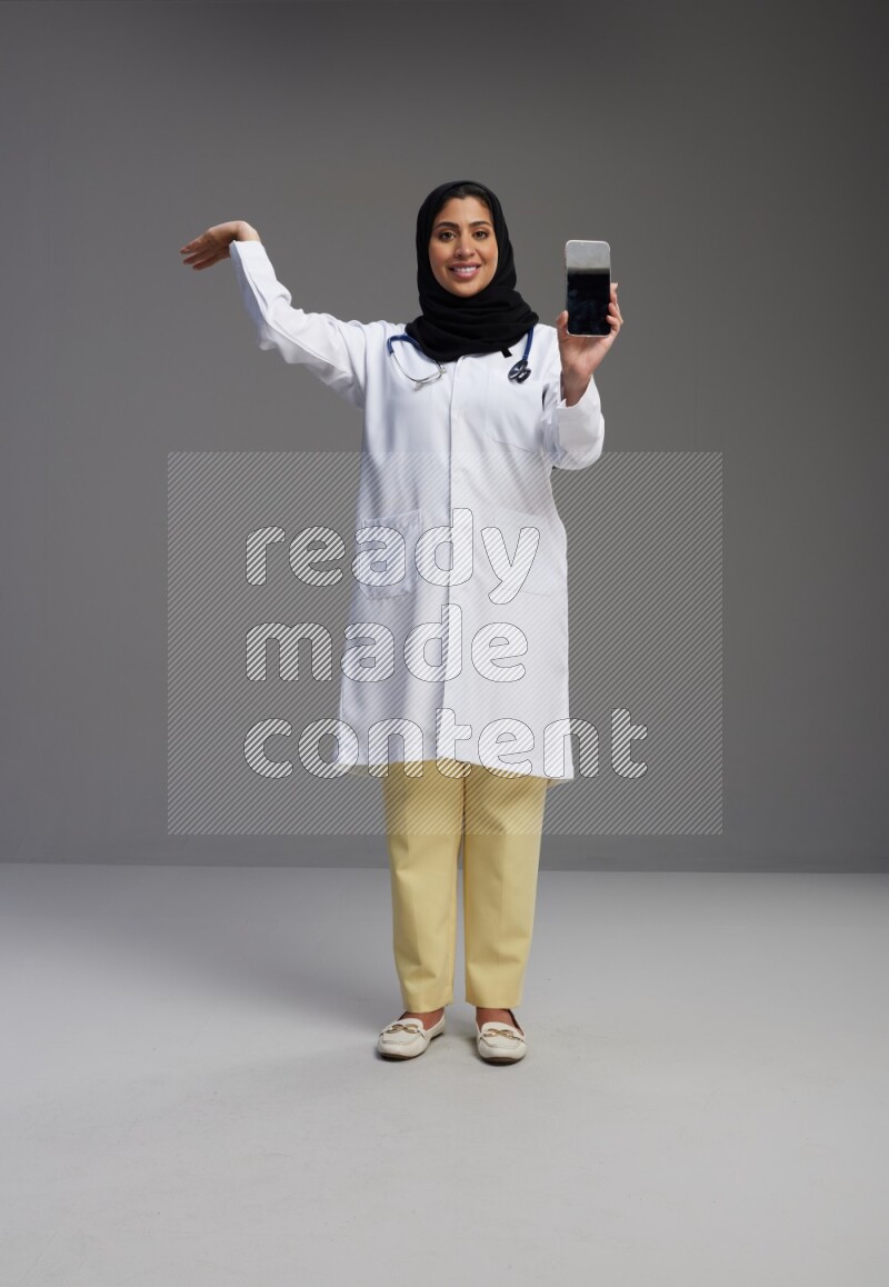 Saudi woman wearing lab coat with stethoscope standing showing phone to camera with sign in the back on Gray background