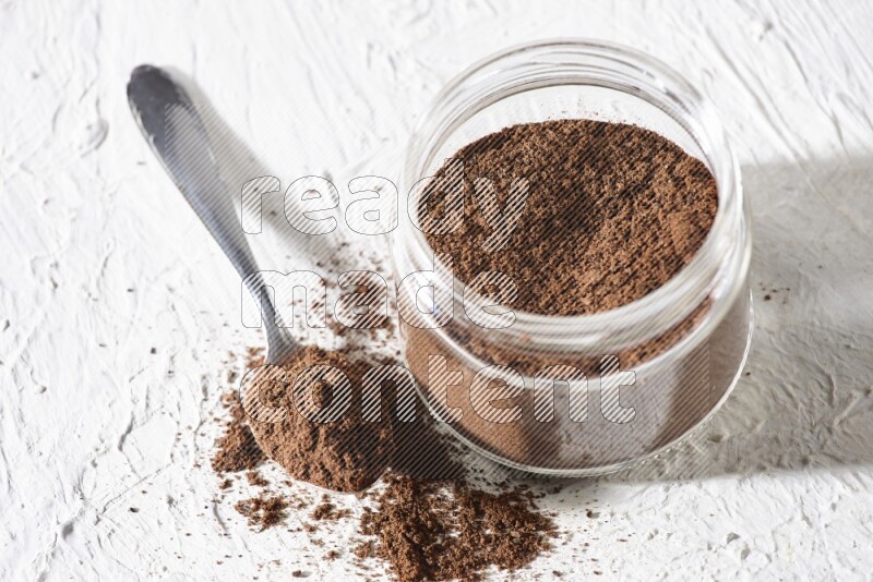 A glass jar full of cloves powder with a metal spoon on a textured white flooring